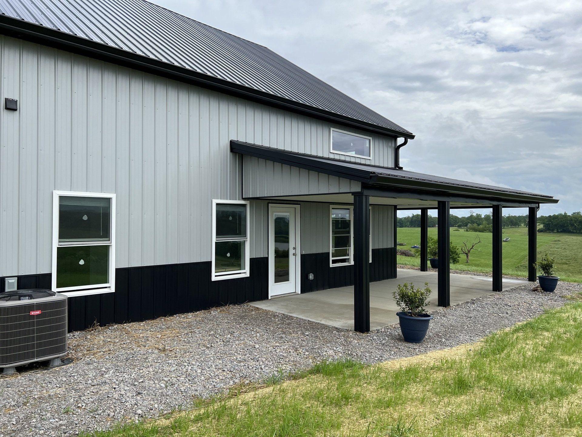 A modern grey and black metal barn-style home with a covered porch, gravel landscaping, and surrounding grassy field.