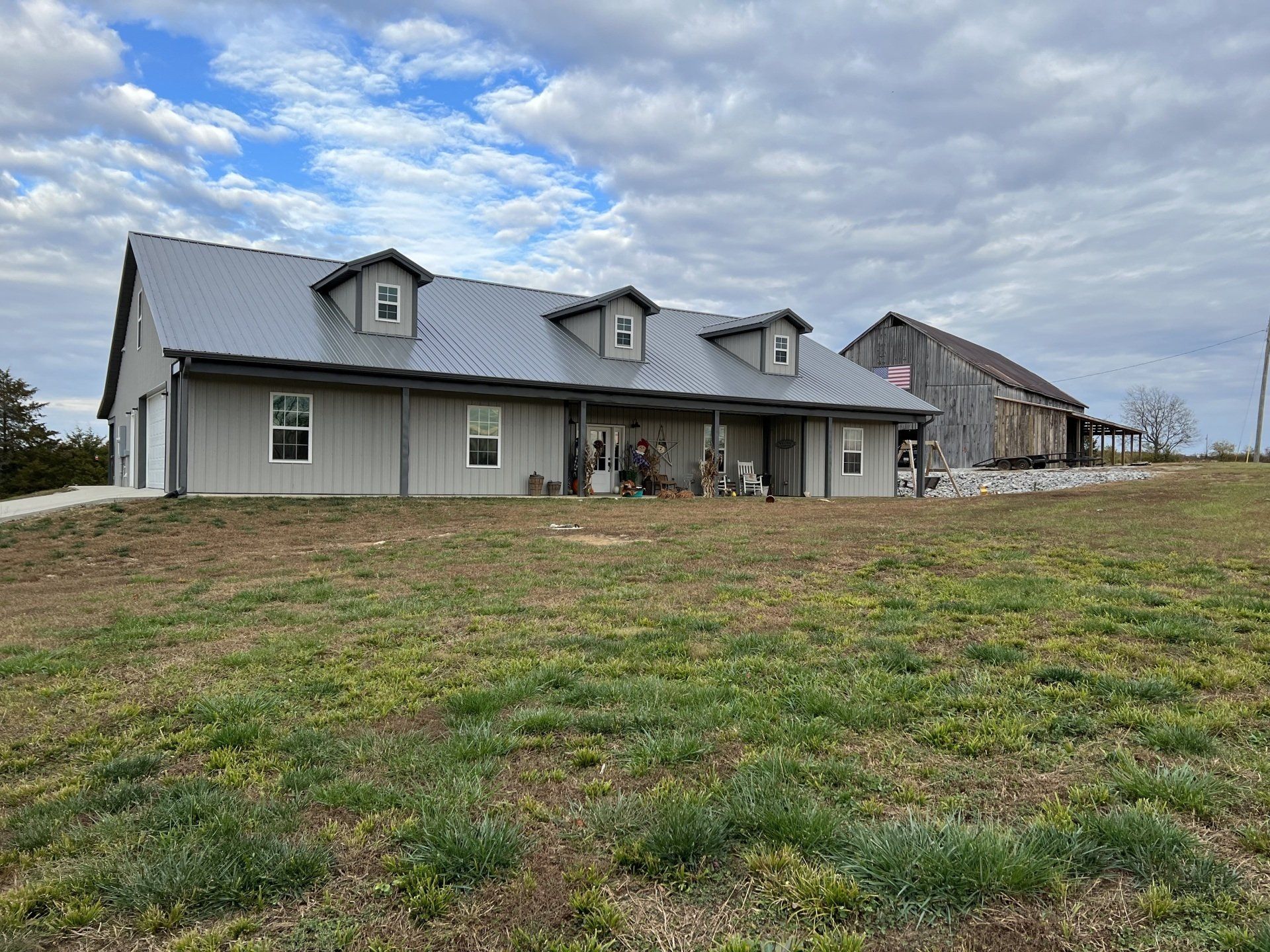 A long, grey metal-sided house with three roof dormers stands in a field next to an old, weathered wooden barn.