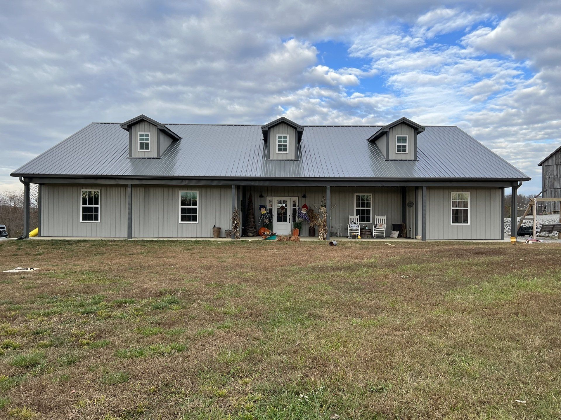 Single-story light gray house with a metal roof and three dormers, featuring a full-length front porch on a grassy lot.