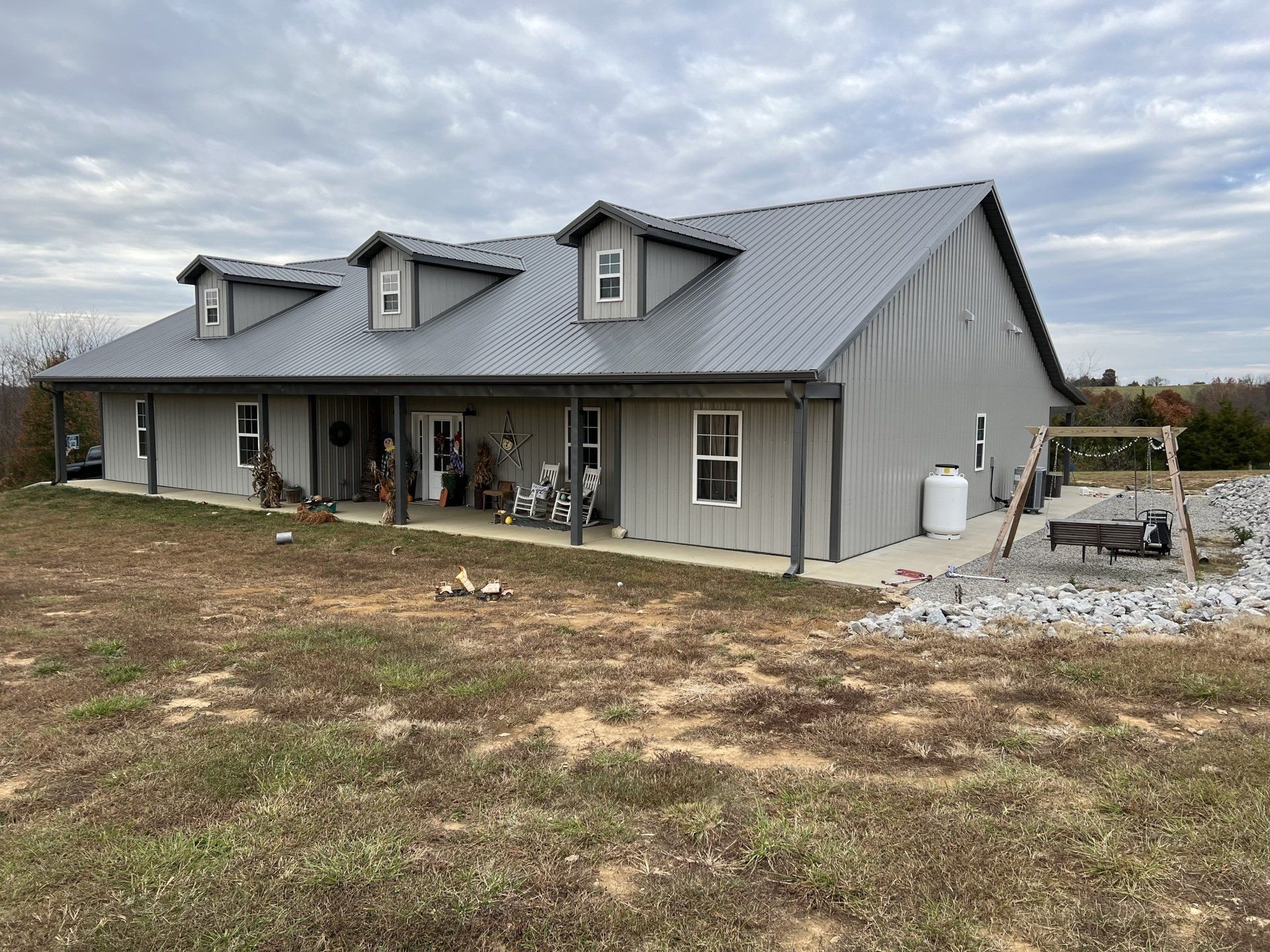 A grey, single-story barndominium-style house with a metal roof, three dormer windows, and a front porch.