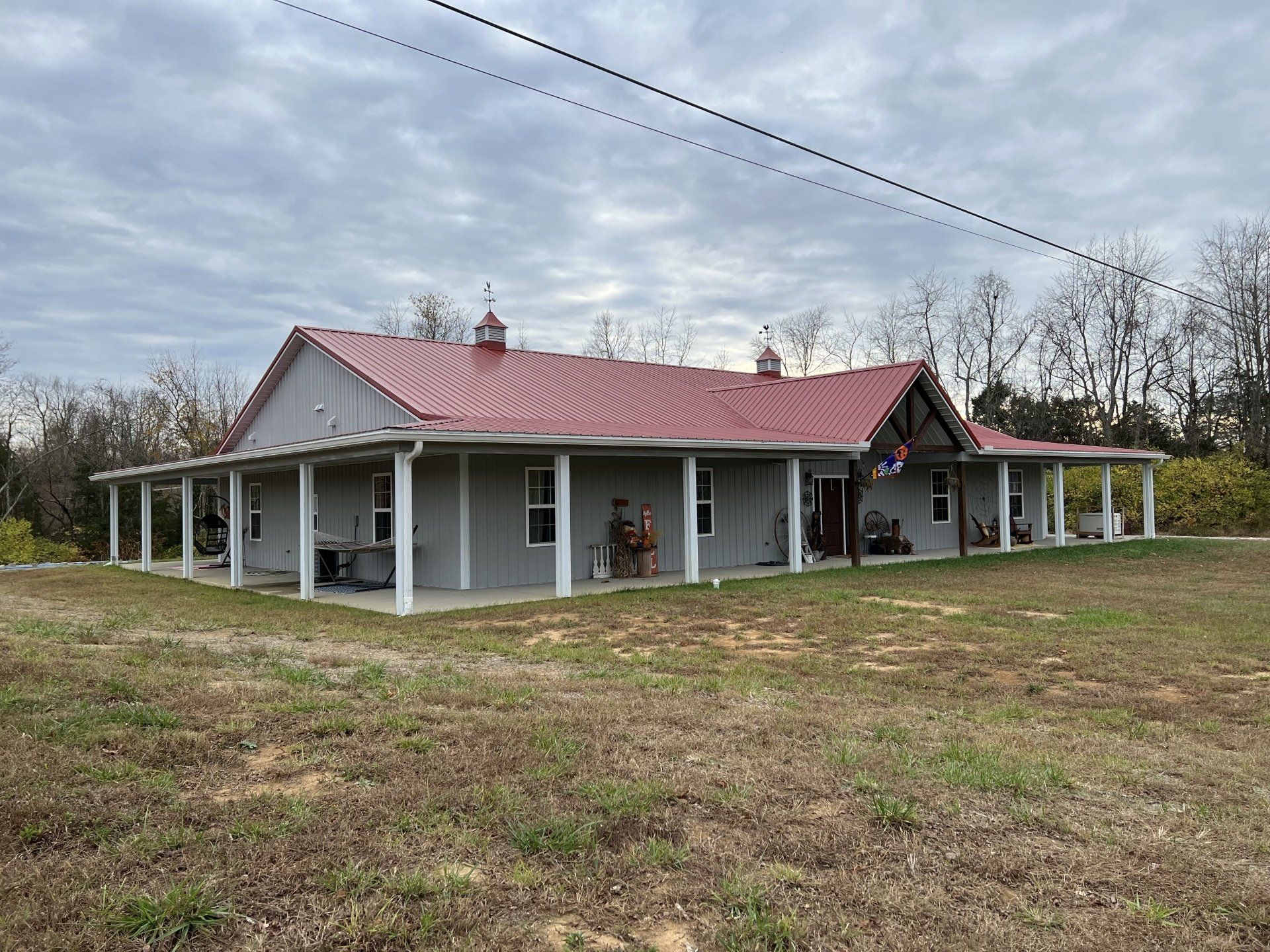 A single-story gray house with a red metal roof and a wrap-around porch, set against a backdrop of trees under a cloudy sky.