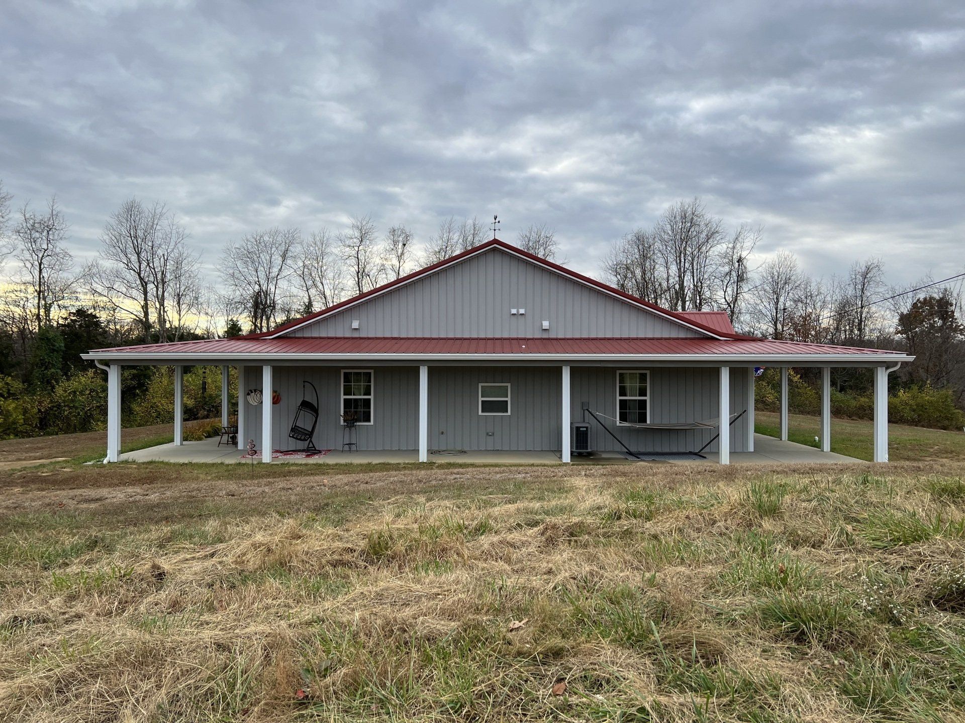 A grey, single-story house with a red metal roof and a wrap-around porch, set against a backdrop of trees under a sky.