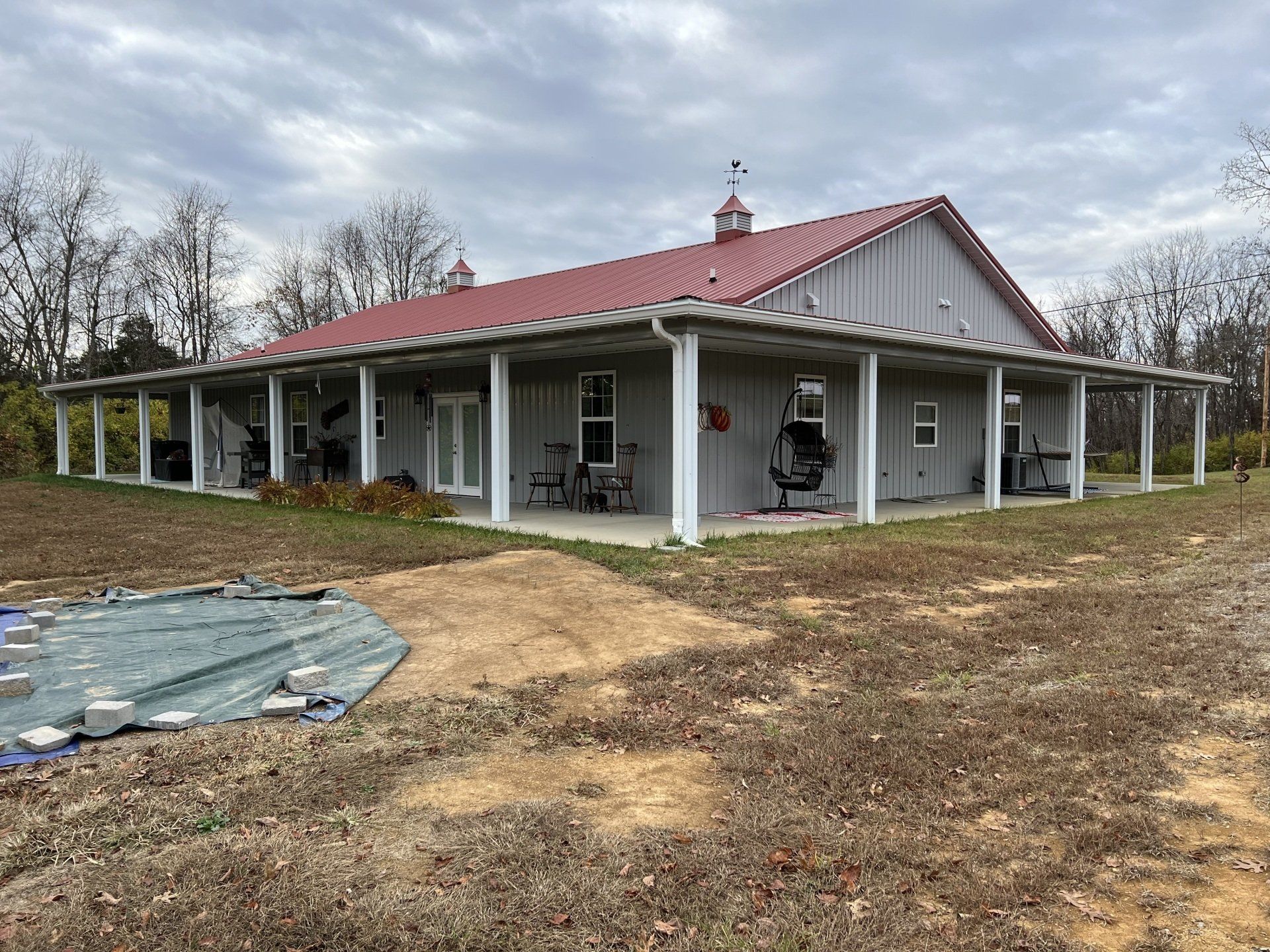 A gray, single-story house with a red metal roof and a wrap-around porch, situated on a dirt lot under a cloudy sky.