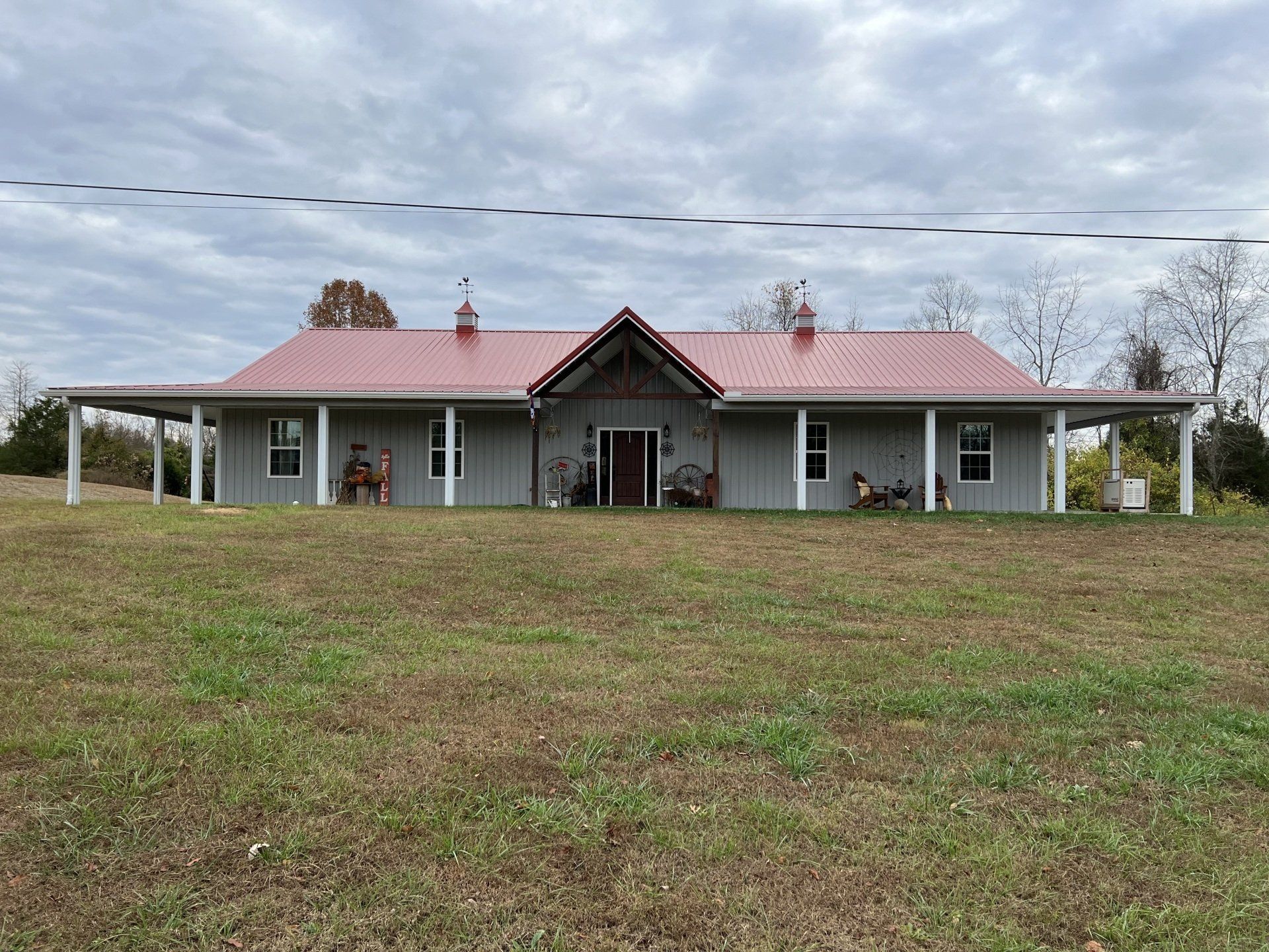 A wide, grey, single-story house with a red metal roof, wraparound porch, and two roof cupolas sits on a grassy hill.
