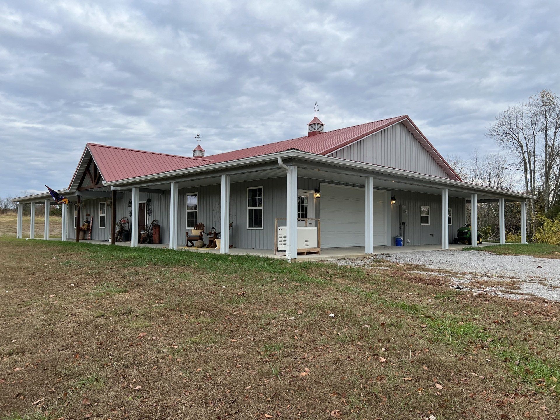 A single-story gray building with a red metal roof and an expansive covered porch, situated on a grassy, open lot.
