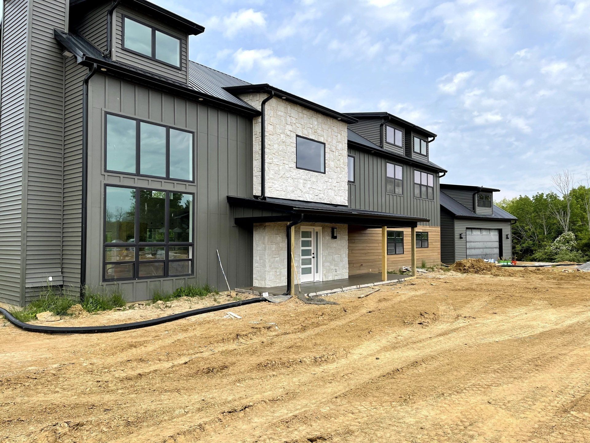A modern, two-story house with dark metal siding and white stone accents under construction on a dirt lot.