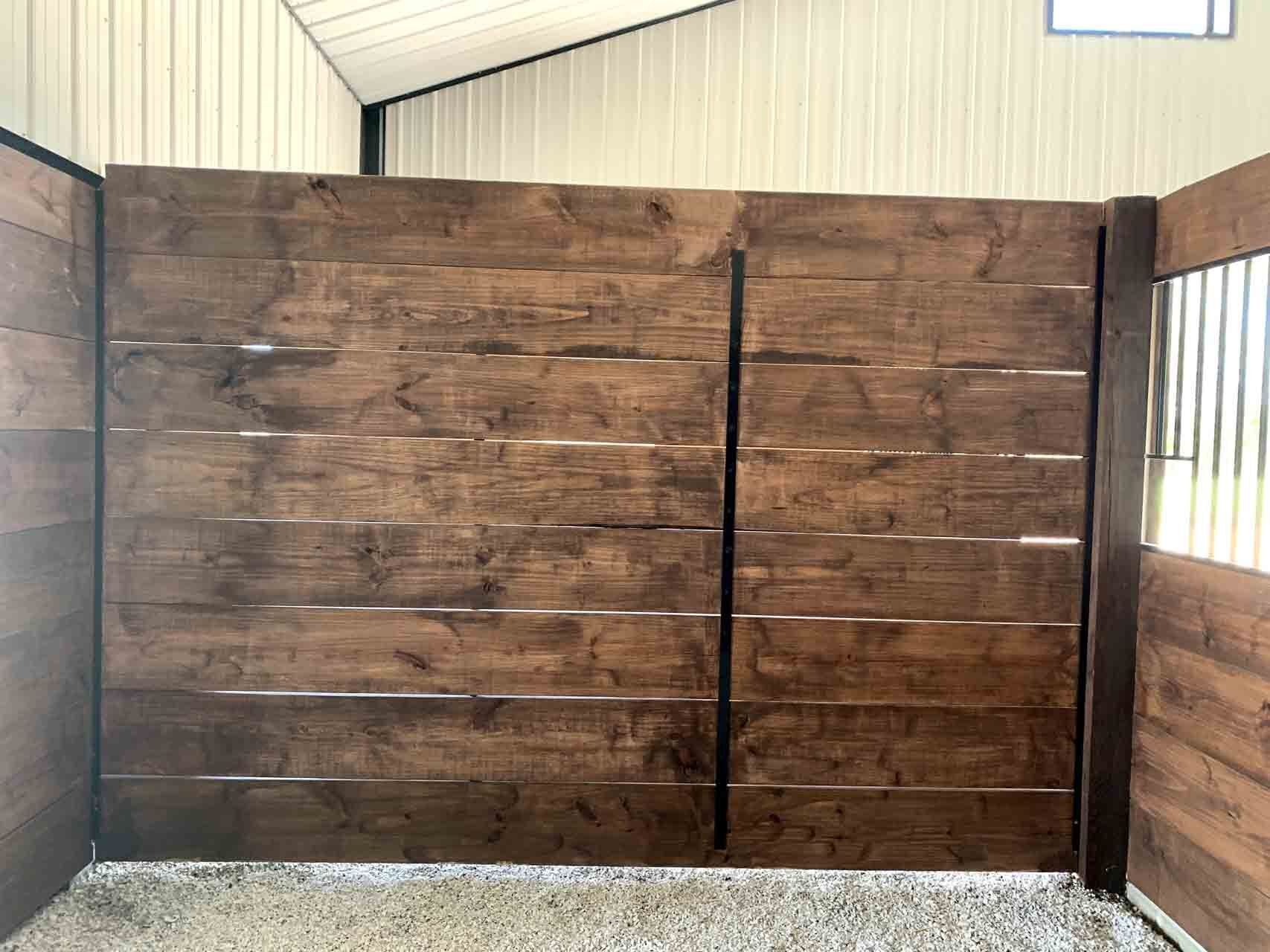 A horse stall with dark-stained horizontal wooden planks, a vertical black frame, and metal bars on the right side.