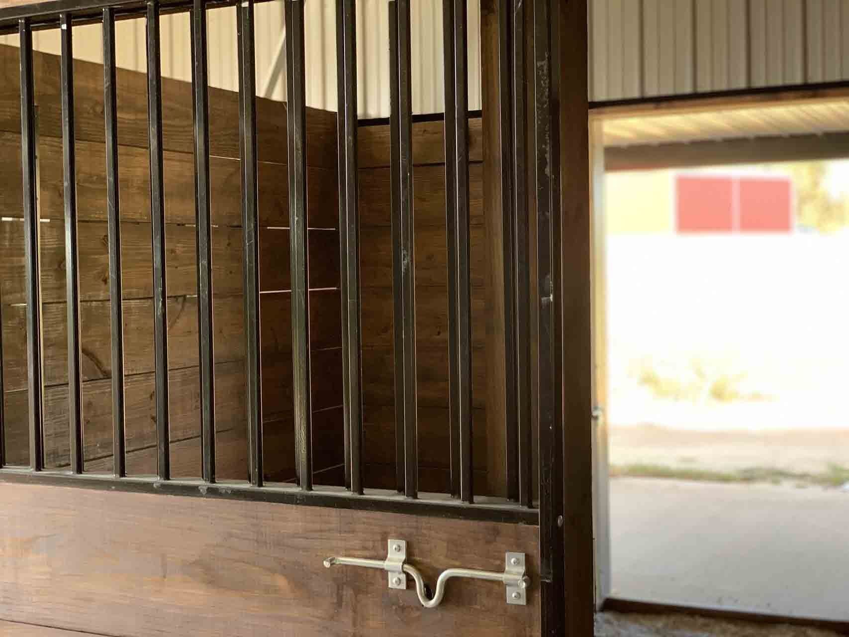 A close-up of a wooden horse stall with vertical black metal bars and a sliding latch, showing an open stable aisle.