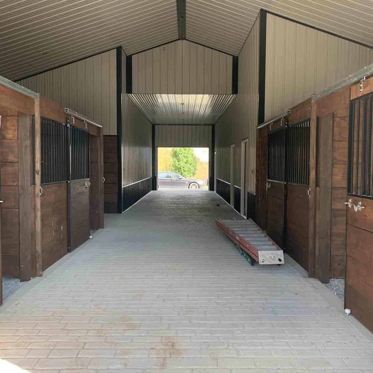 An indoor barn aisle with wooden horse stalls on both sides and a light-colored paved floor leading to an outdoor exit.