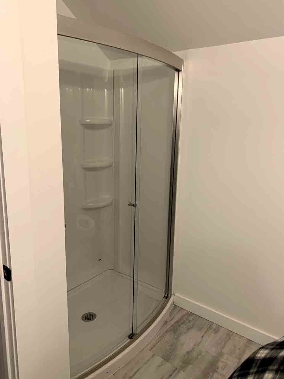 A modern corner shower stall with a curved glass door and white walls set against wood-look flooring.