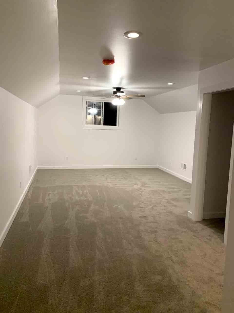 An empty attic room with white walls, slanted ceilings, gray carpet, a ceiling fan, and a single window.