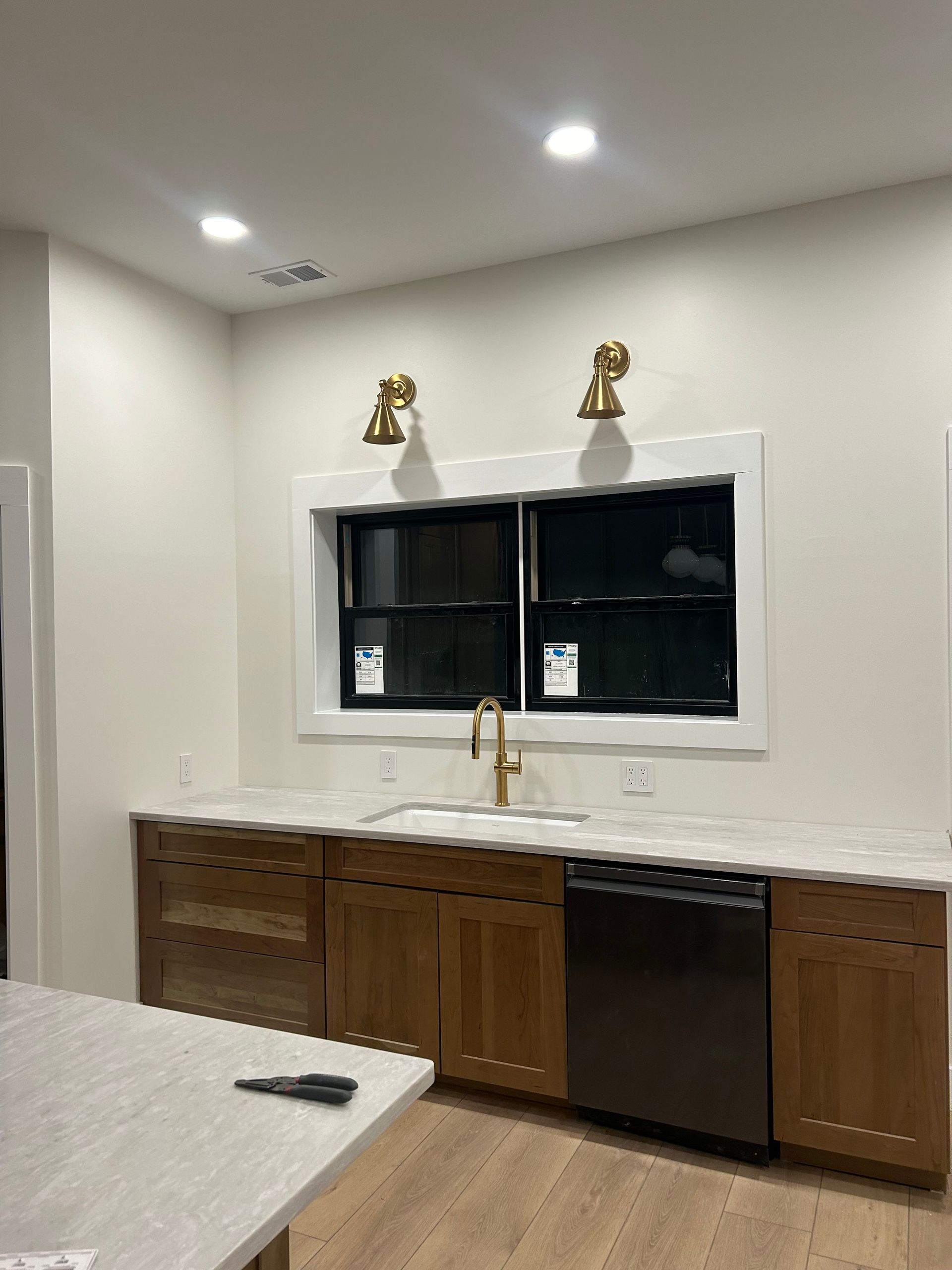 A kitchen view featuring wood cabinets, a black dishwasher, a white countertop, and a window with two brass wall lights.