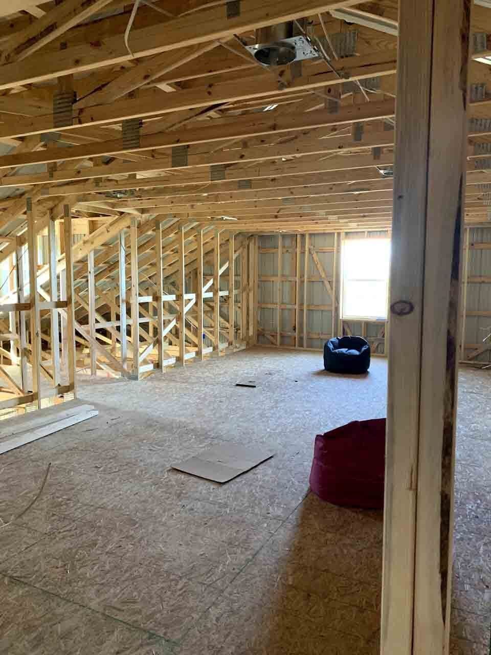 An unfinished attic room featuring exposed wooden rafters, plywood subflooring, a bright window, and two beanbag chairs.