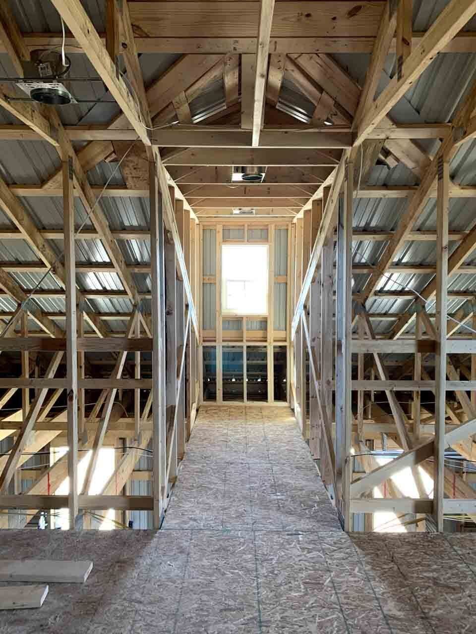 A perspective view of a wooden attic under construction, featuring exposed timber framing and a bright window at the end.