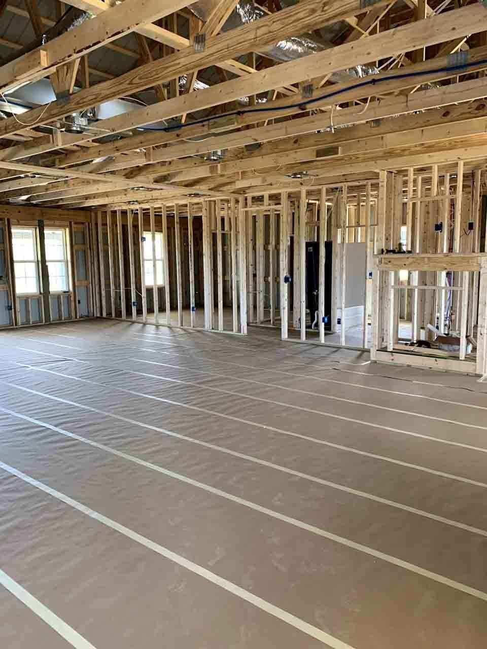 An interior view of a room under construction, showing wooden framing, uncovered ceiling joists, and protective floor paper.