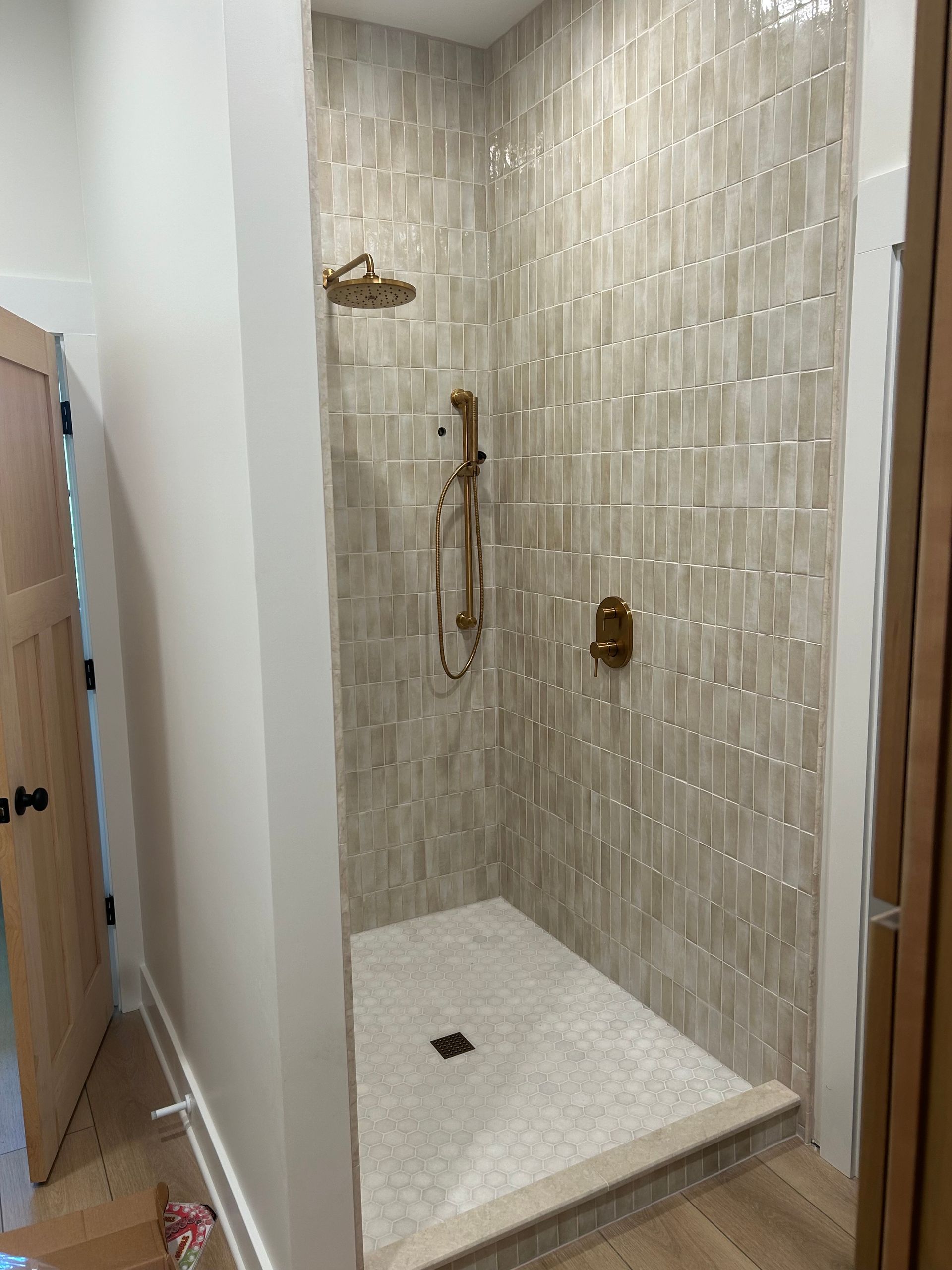 A walk-in shower with vertical, beige-toned rectangular wall tiles, a mosaic tile floor, and brushed-gold fixtures.