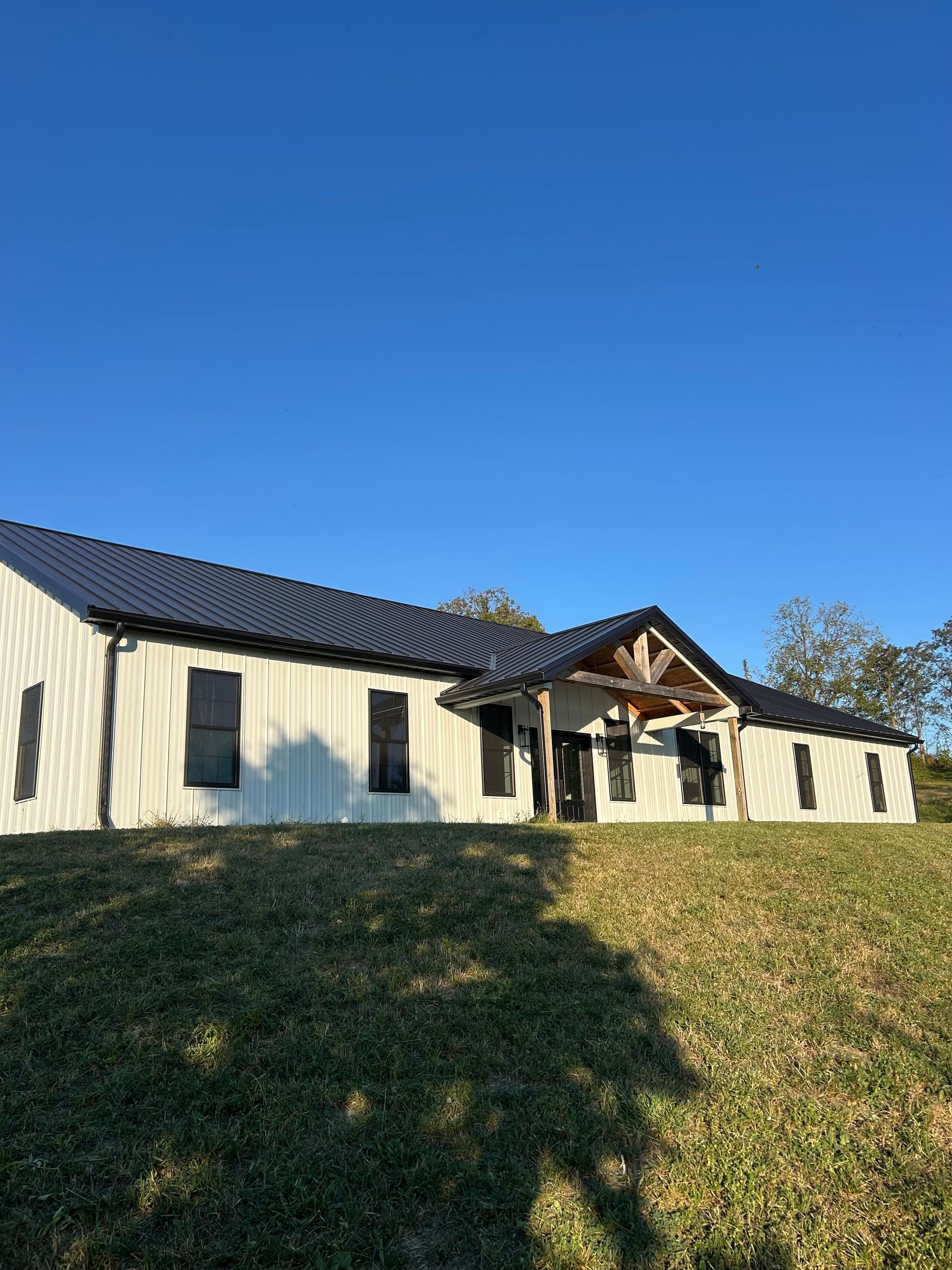A white farmhouse-style home with a dark metal roof and a wooden entryway porch on a grassy hill under a clear blue sky.