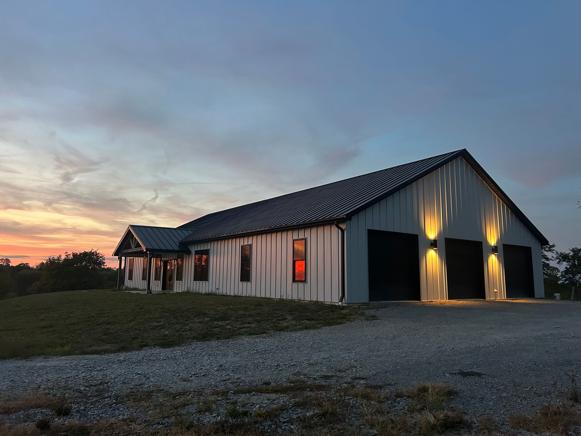 A white, modern pole barn home with a dark metal roof and three garage bays, illuminated at sunset.