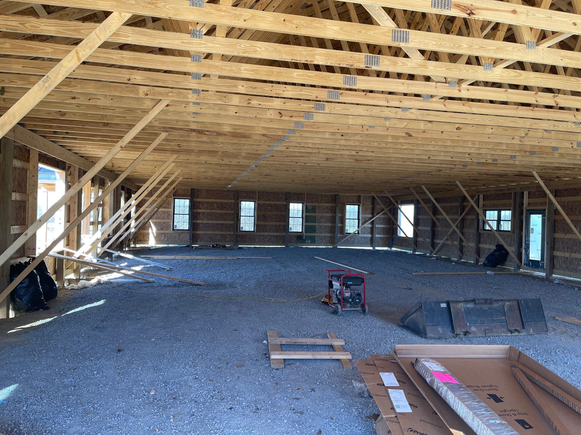 Interior view of a large unfinished building with exposed wooden roof trusses, gravel flooring, and several wall windows.