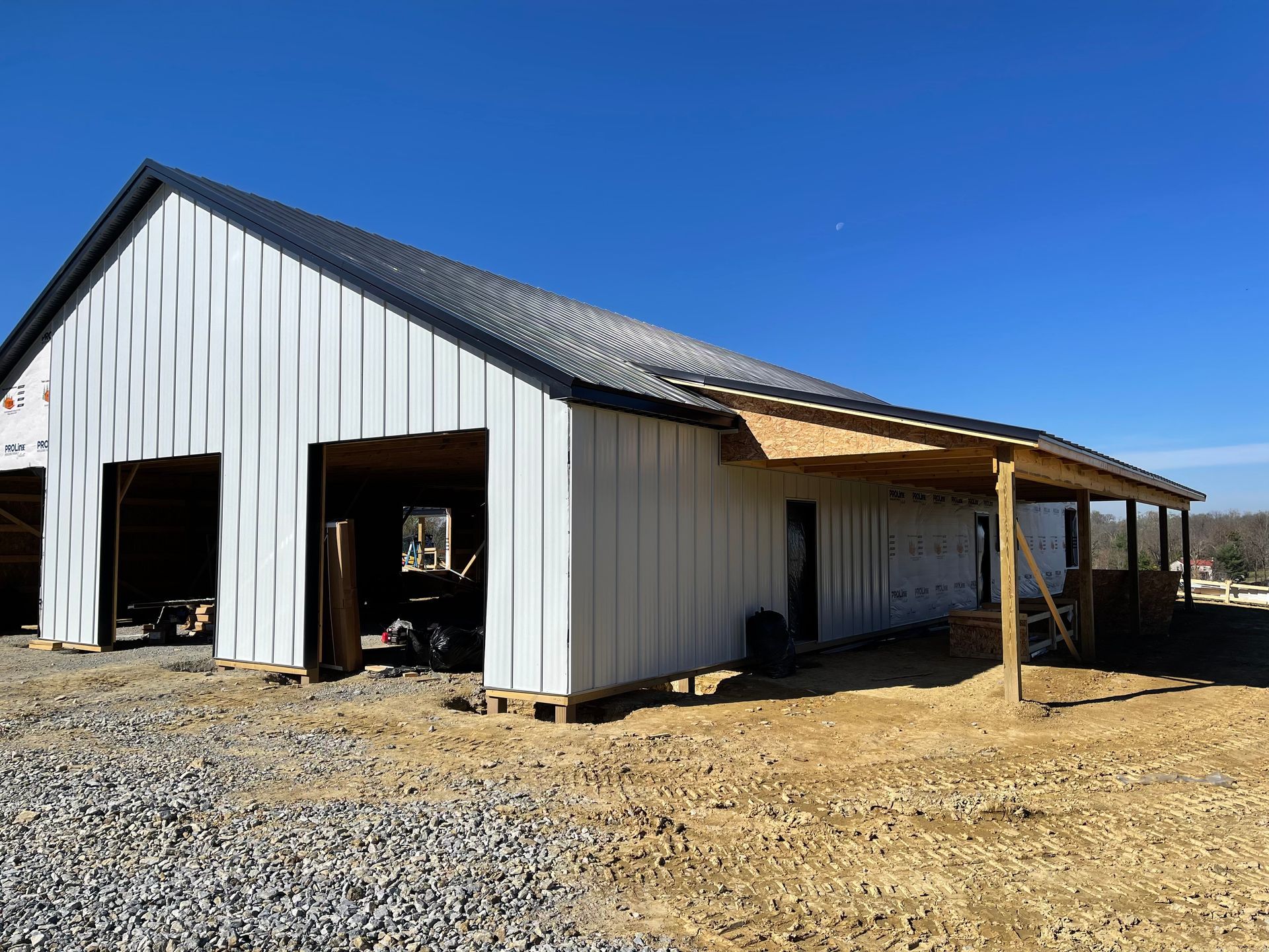 A white metal-sided building under construction with an open doorway and a side porch, set against a clear blue sky.