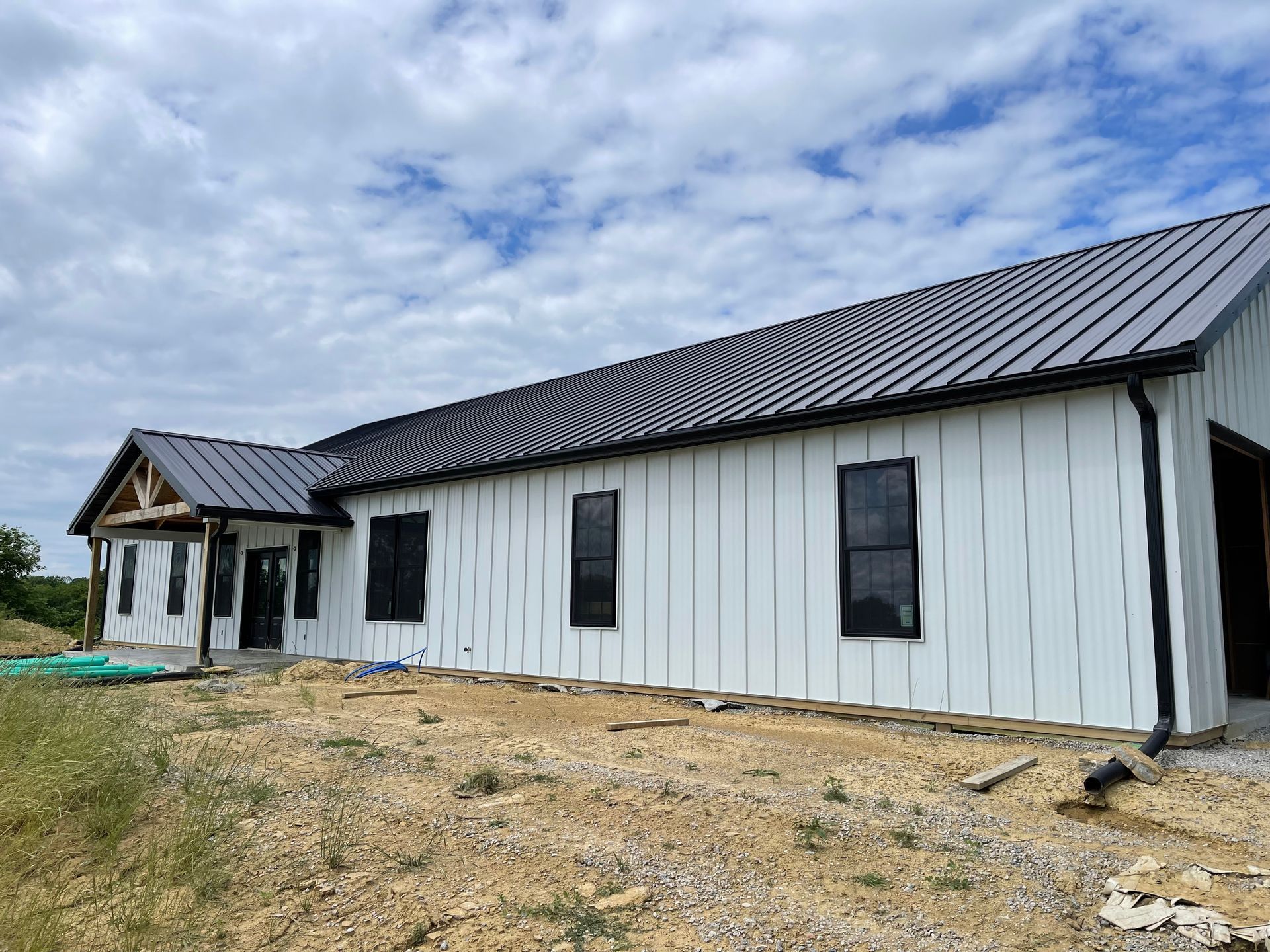 A new house under construction featuring white vertical siding, a dark metal roof, black window frames, and a porch.