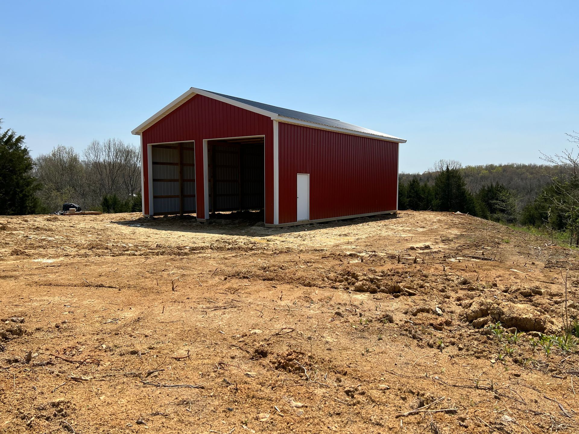 A red metal barn with two open garage bays sits on a cleared, dirt lot under a clear blue sky.