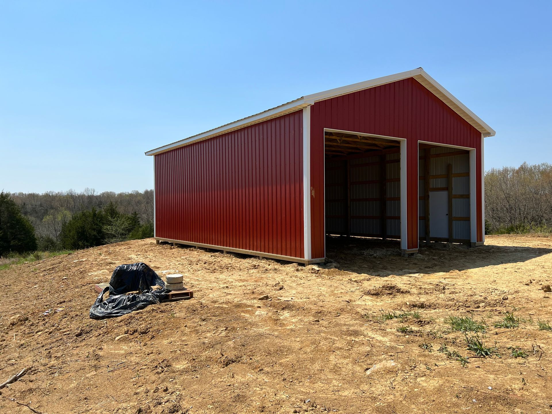A new red metal pole barn stands on a cleared, dirt-covered hill under a clear blue sky.