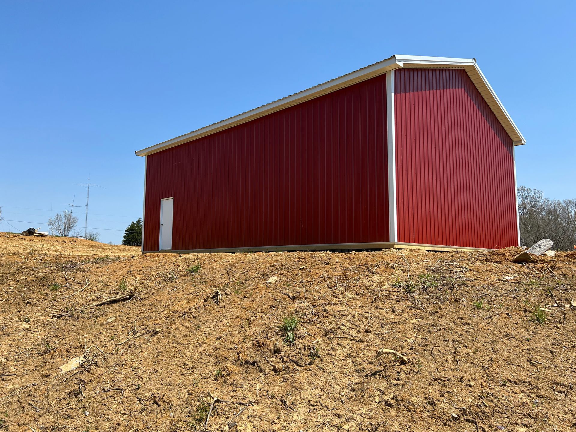 A red metal barn with a white trim and door sits on a dirt-covered slope under a clear blue sky.