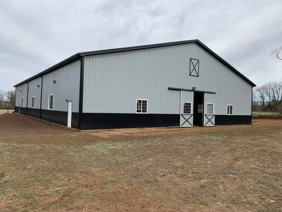 A modern, gray metal barn with black trim and sliding front doors, set on a grassy lot under a cloudy sky.