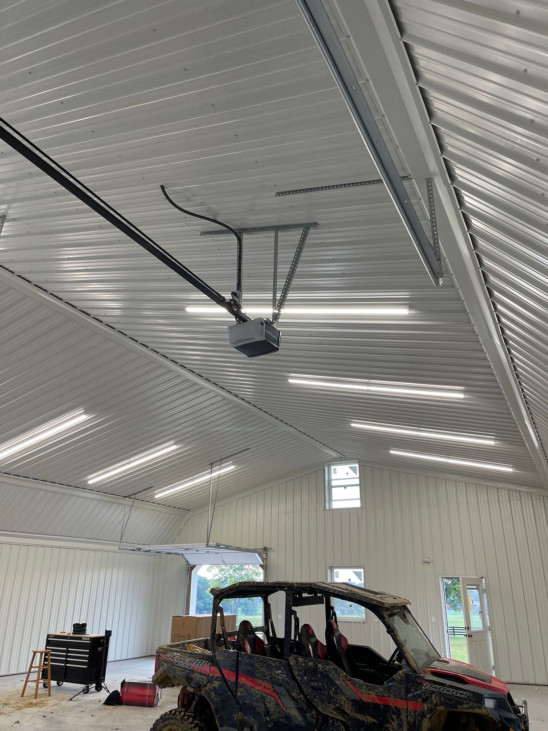 A high-ceilinged workshop with corrugated metal walls, a ceiling-mounted garage door opener, and a muddy utility vehicle.