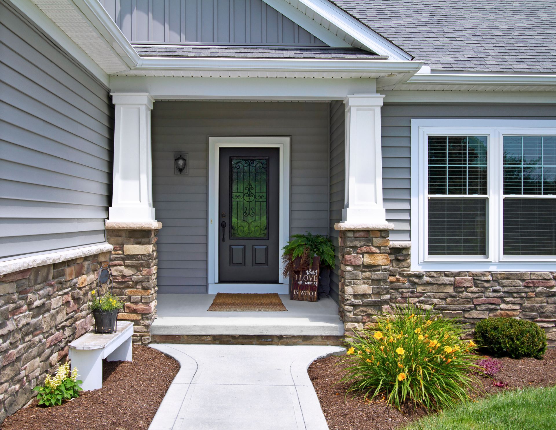 A house entryway with gray horizontal siding, stone veneer base, two white pillars, a dark front door, and a walkway.