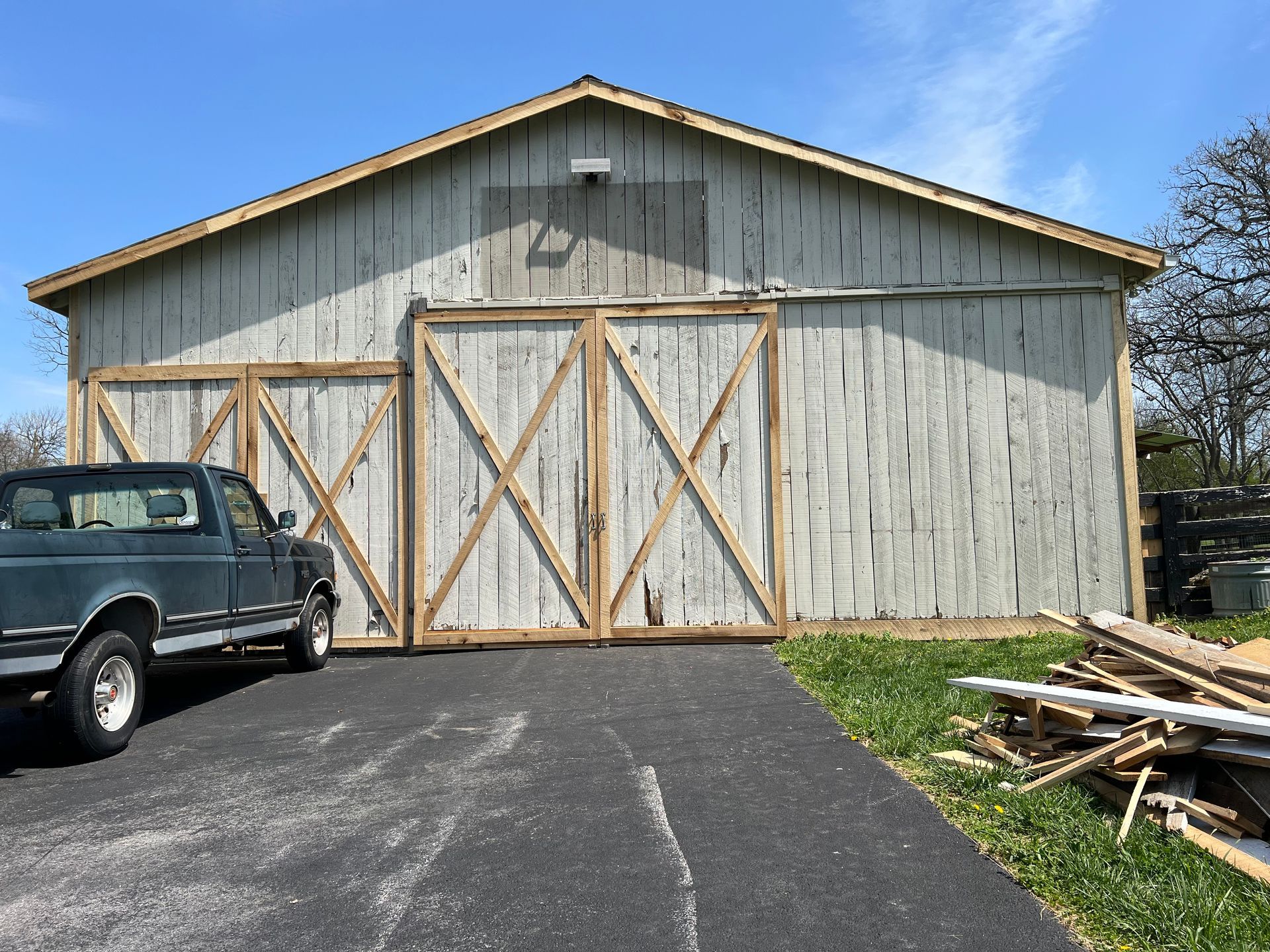 A weathered barn with sliding wooden doors parked next to a dark pickup truck on an asphalt driveway.