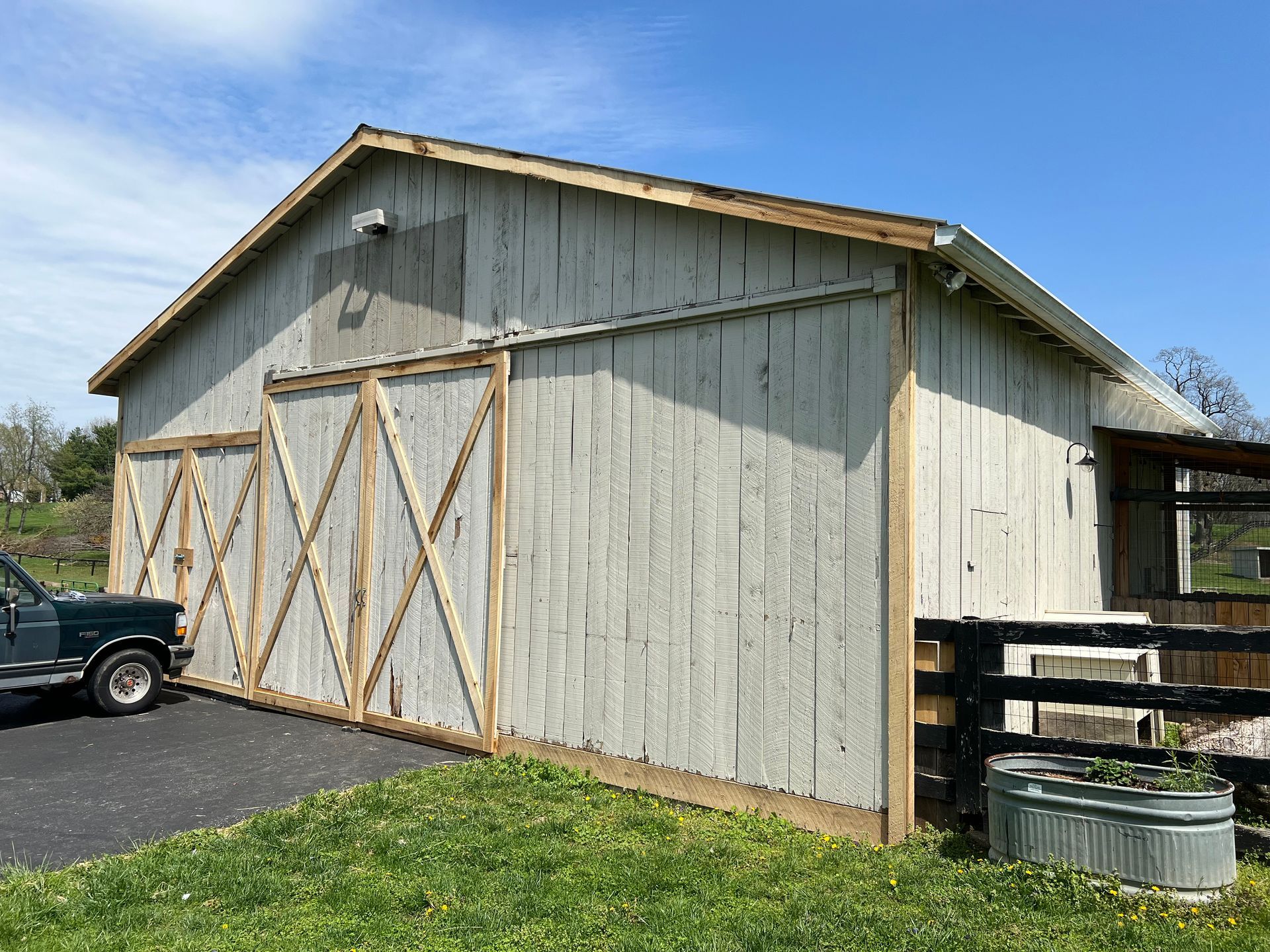 A weathered, light-colored barn with sliding wooden doors, an adjacent black fence, and a metal trough in a grassy yard.