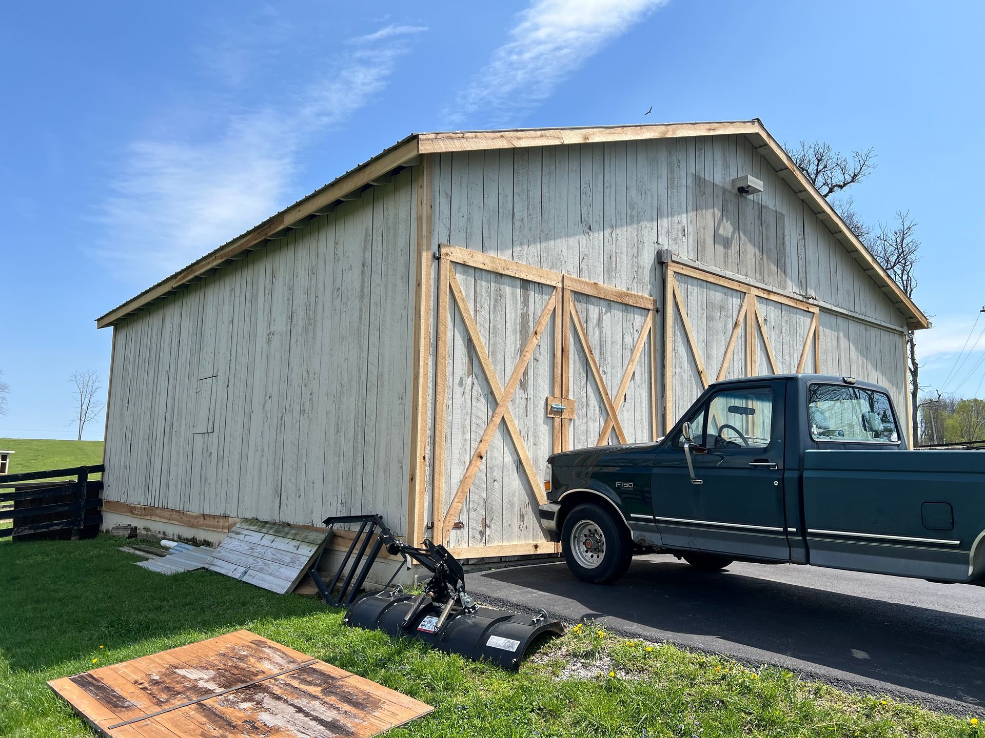 A dark green pickup truck parked on an asphalt driveway next to a light-colored wooden barn with double cross-brace doors.