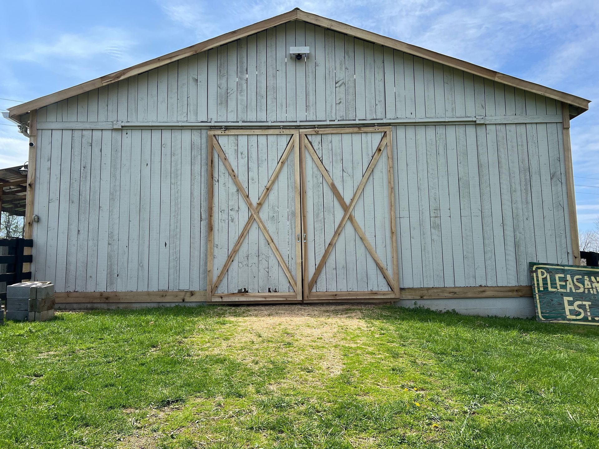 A weathered white barn with large, X-braced sliding doors stands behind a patch of green grass under a blue sky.