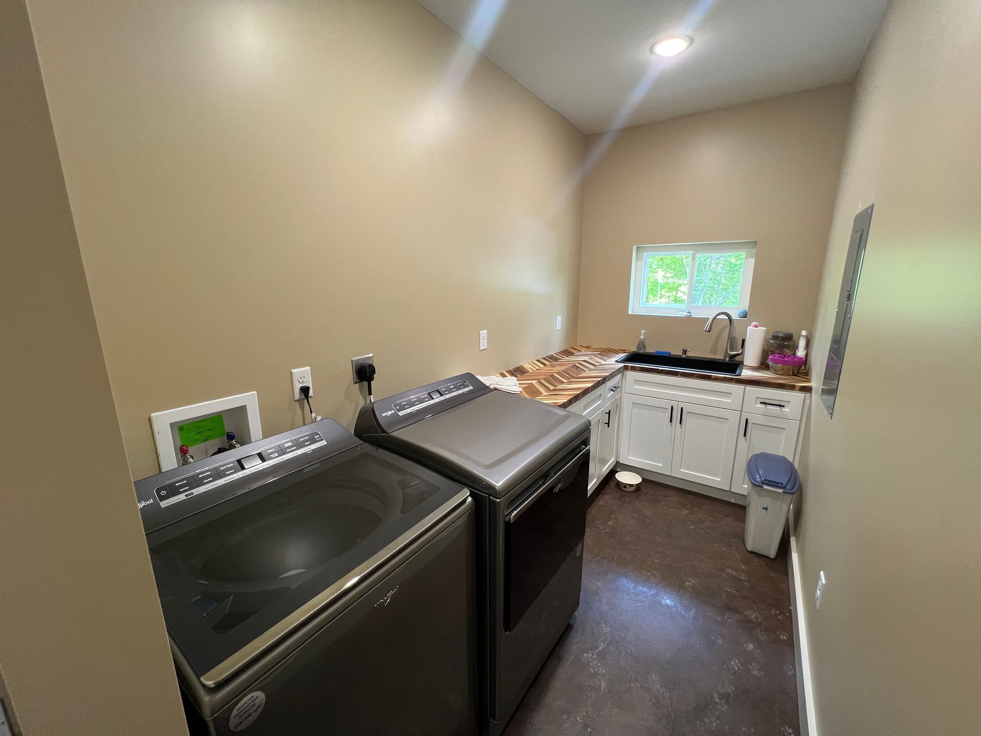 Laundry room with a grey washer and dryer, white cabinets, a sink, and a window against light tan walls.