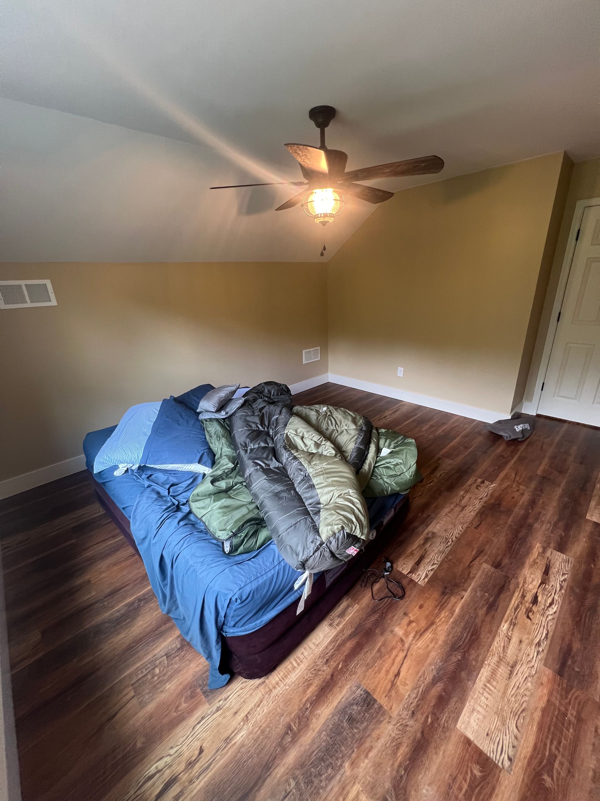 A bed with blue bedding and stacked sleeping bags in a room with wood-look flooring, tan walls, and a ceiling fan.