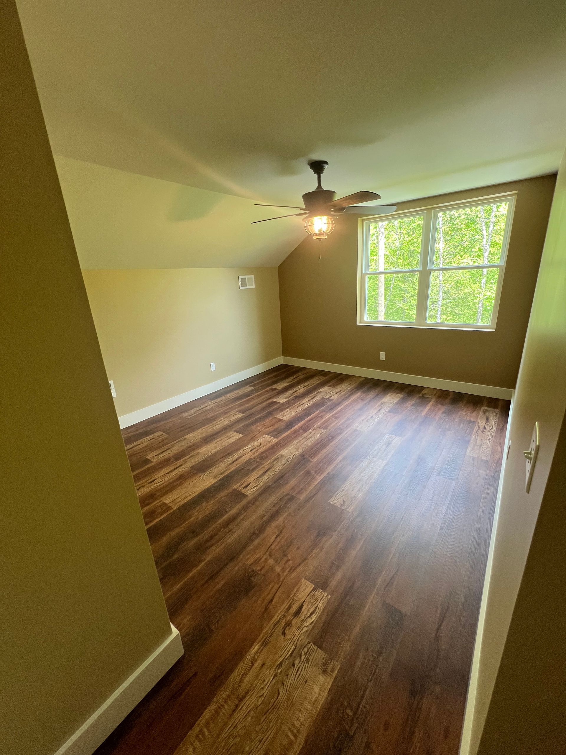 Empty room with wood-look flooring, sloped ceiling, ceiling fan, and a large window looking out onto green trees.