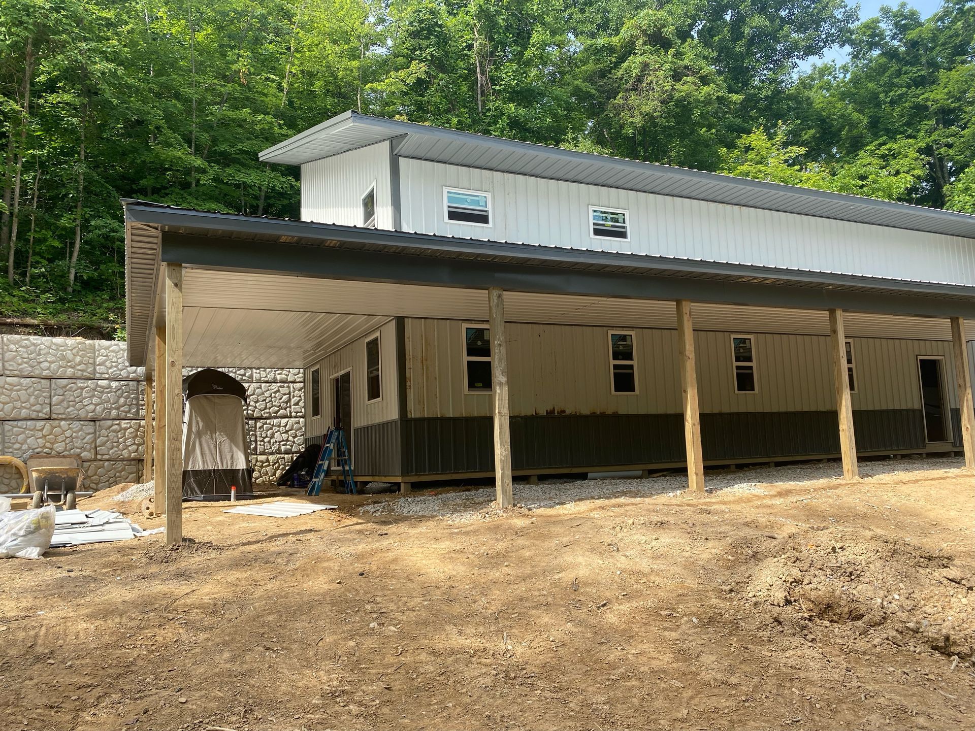 A white and gray metal building with a shed-style roof and a long covered porch, situated on a dirt lot near a forest.