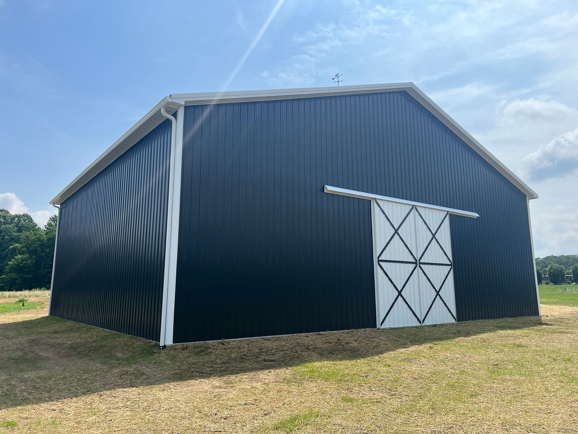 A black pole barn with a white sliding door and white trim set in a grassy field under a sunny blue sky.