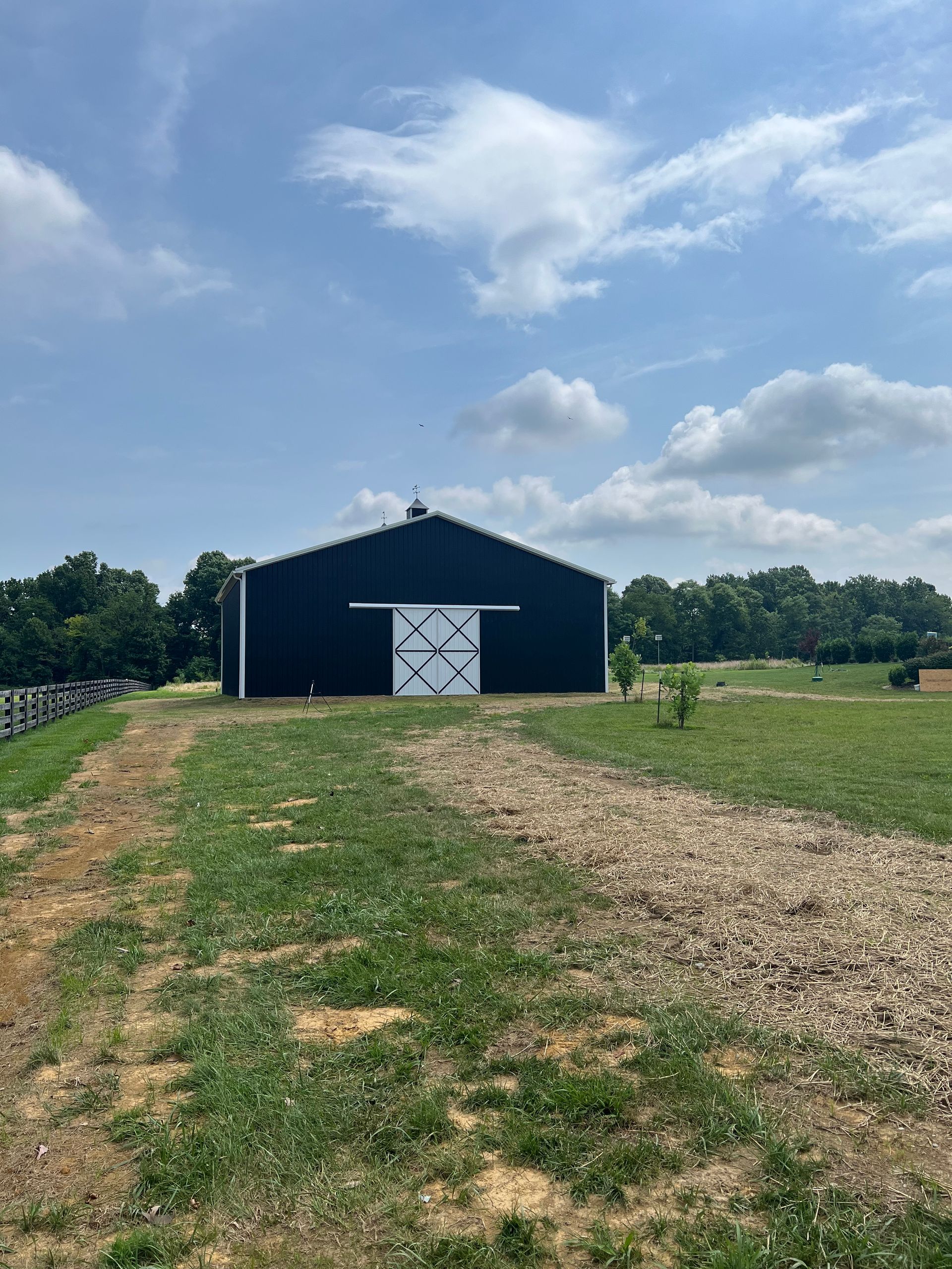 A black barn with white sliding doors sits in a grassy field under a bright blue sky with scattered clouds.