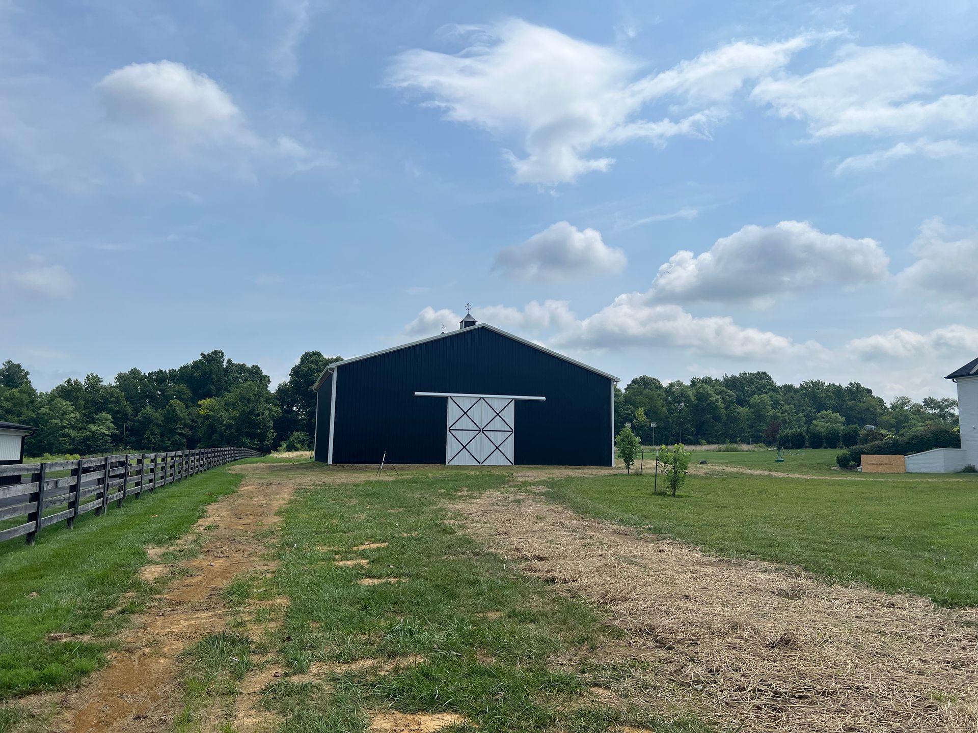 A black barn with white sliding doors sits in a grassy field under a blue sky with white clouds.