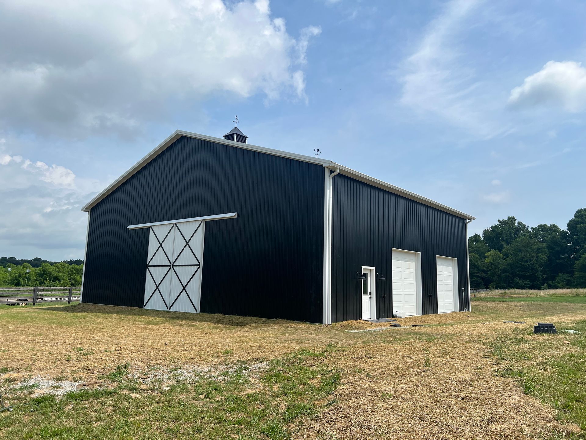 A black barn with white sliding doors and trim stands on a grassy field under a bright, cloudy blue sky.