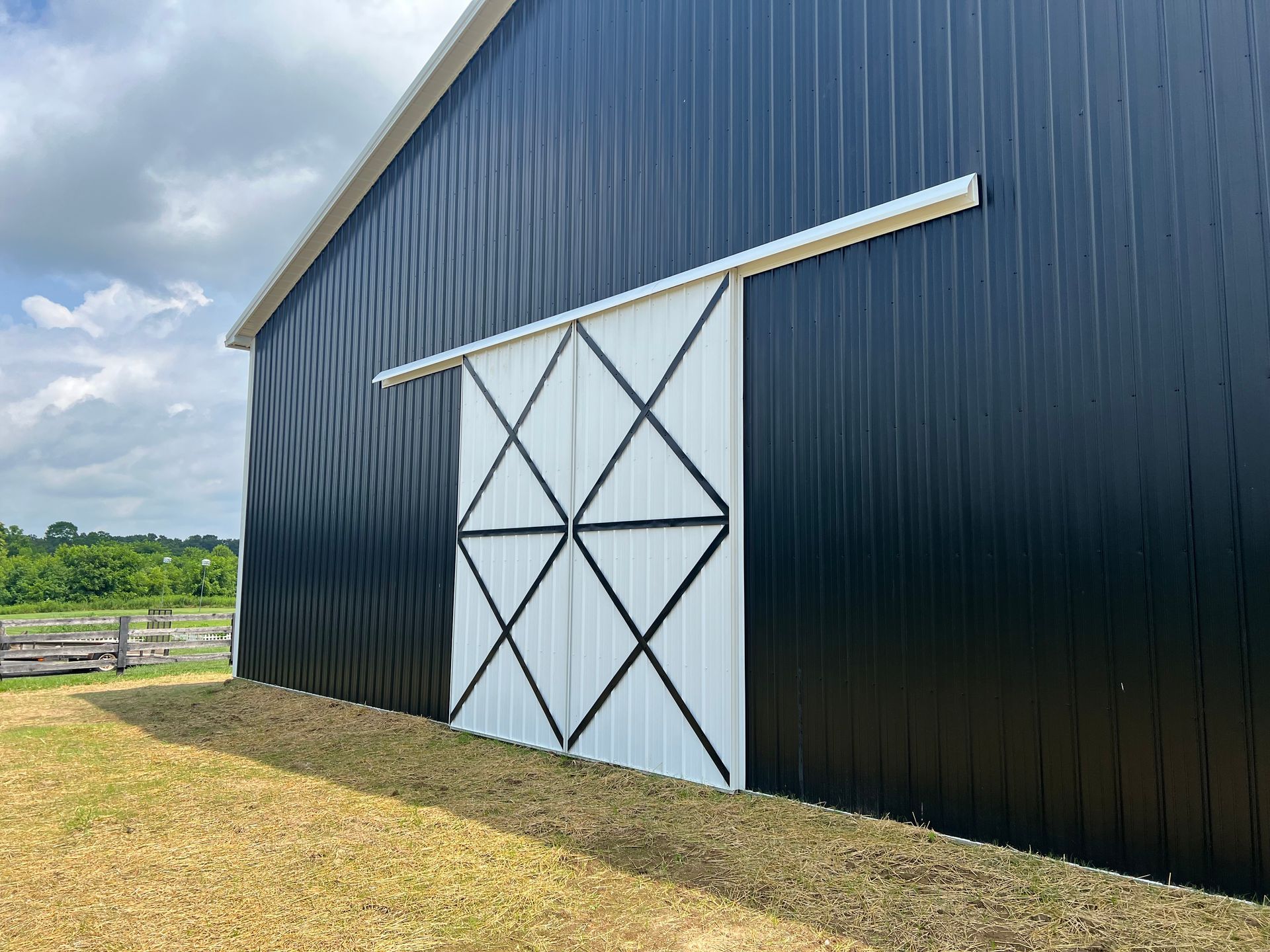 A large black barn featuring a sliding white door with black diagonal trim, set in a grassy field under a cloudy sky.