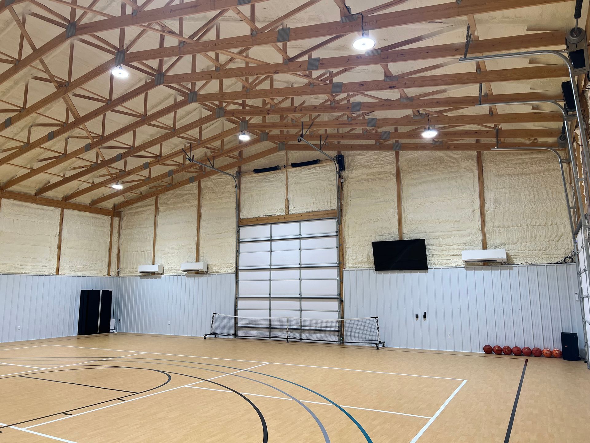 A modern indoor sports court with a basketball floor, high vaulted wooden ceiling, overhead lights, and metal walls.
