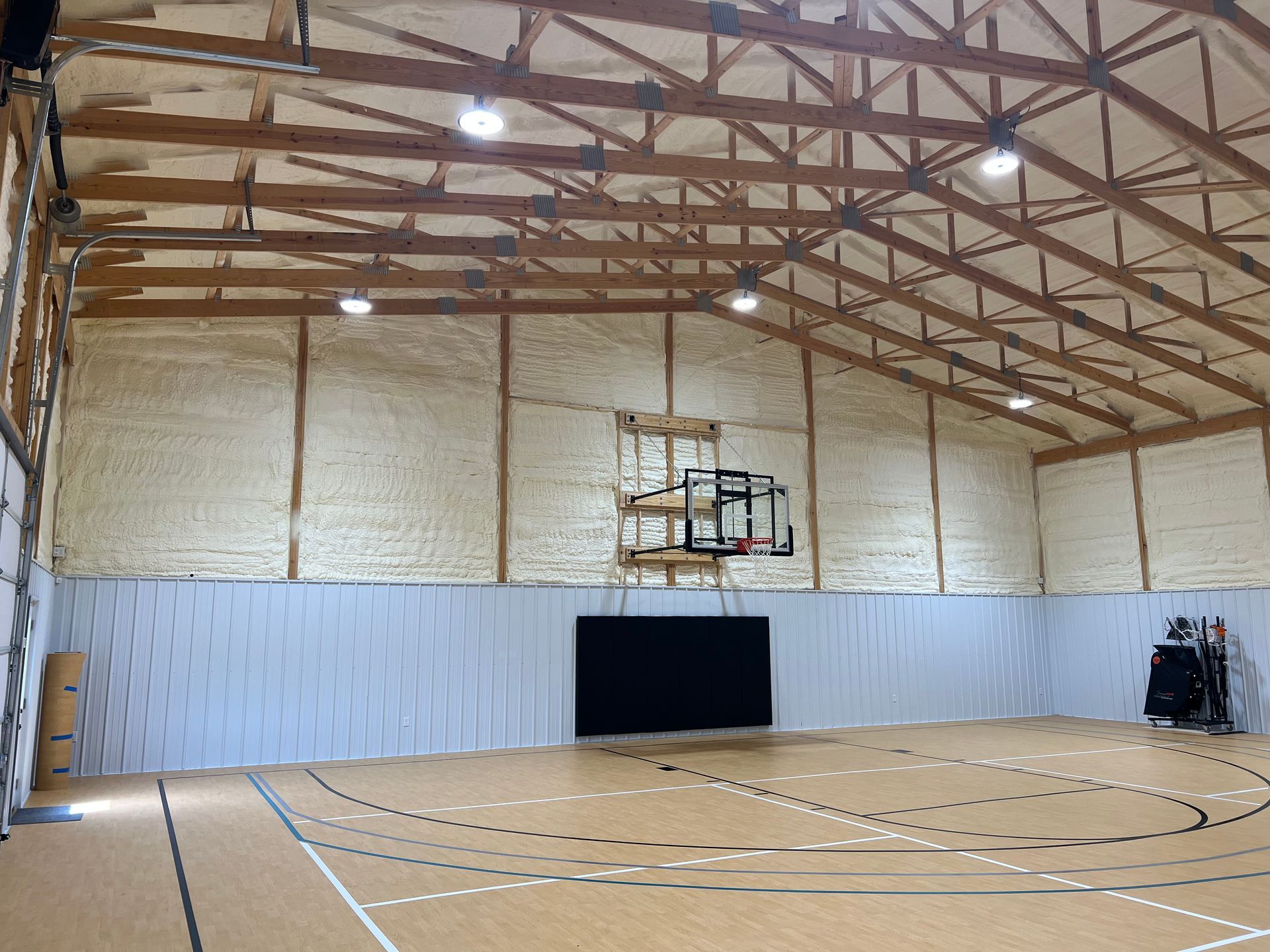 Indoor basketball court with wooden floors, white insulated walls, a hoop, and overhead lighting.