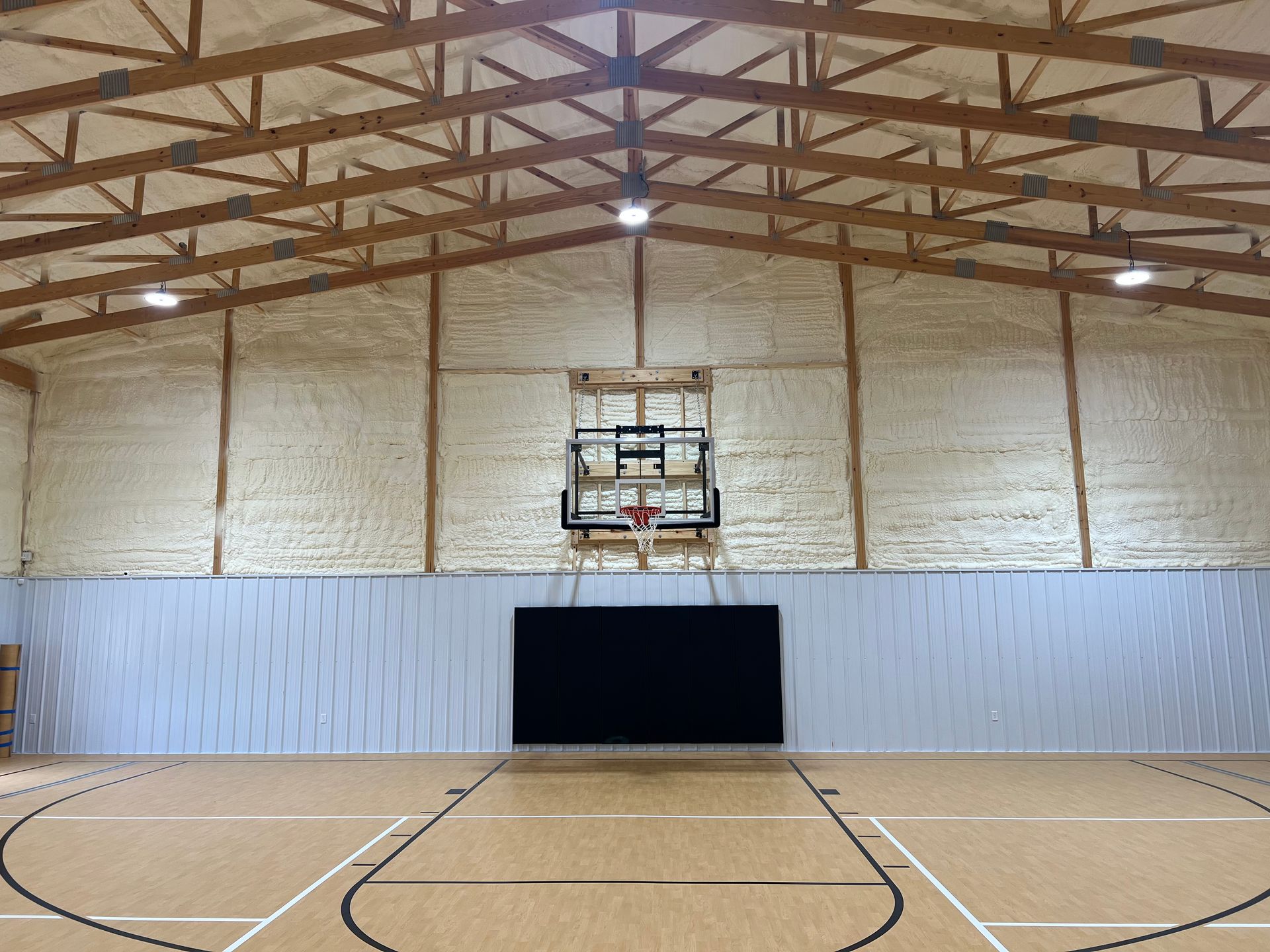 A brightly lit indoor basketball court with a tan floor, white walls, exposed wooden trusses, and a basketball hoop.