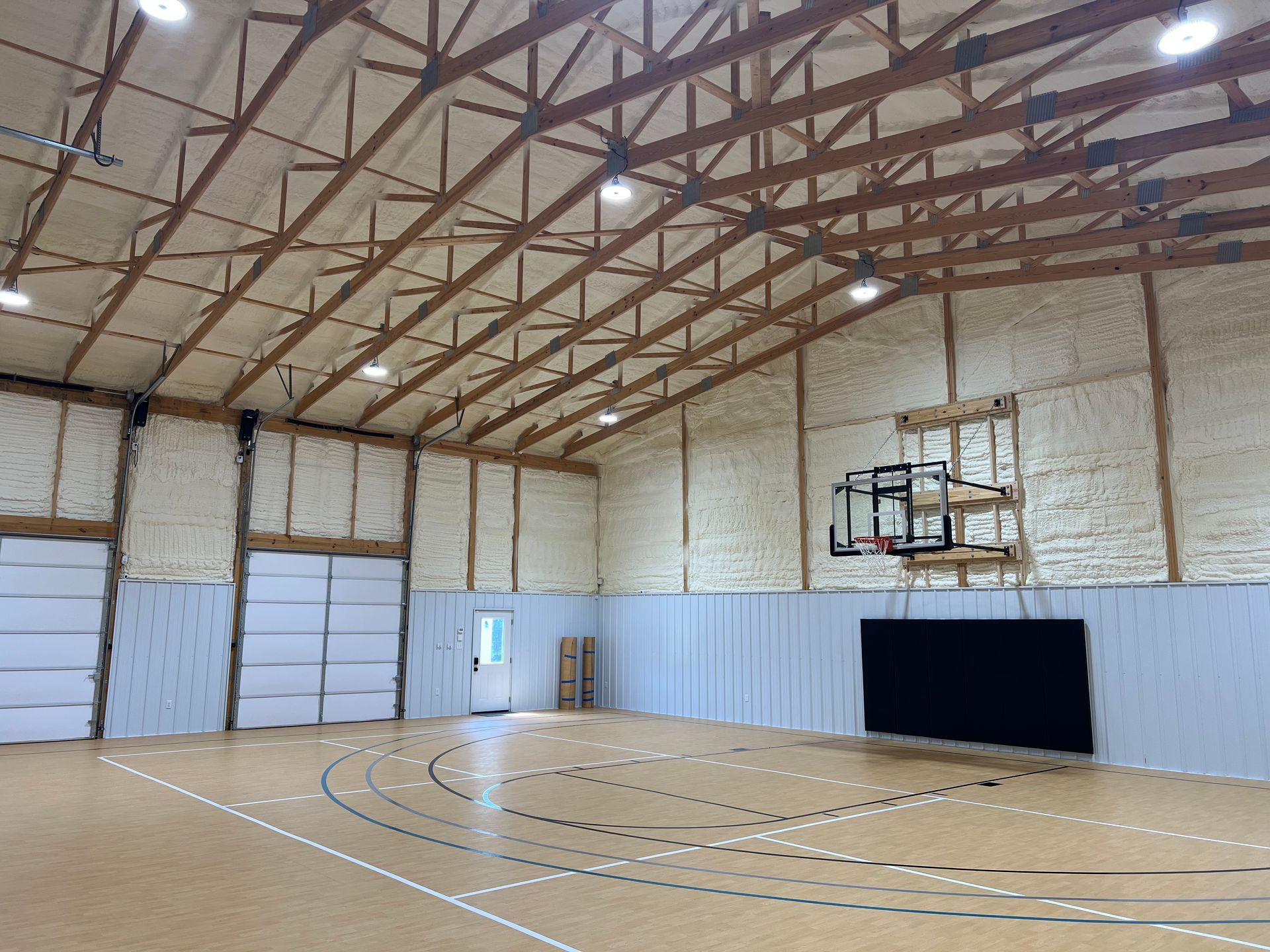 Indoor basketball court with a hoop, a wooden floor, and spray foam insulation on the walls and vaulted ceiling.