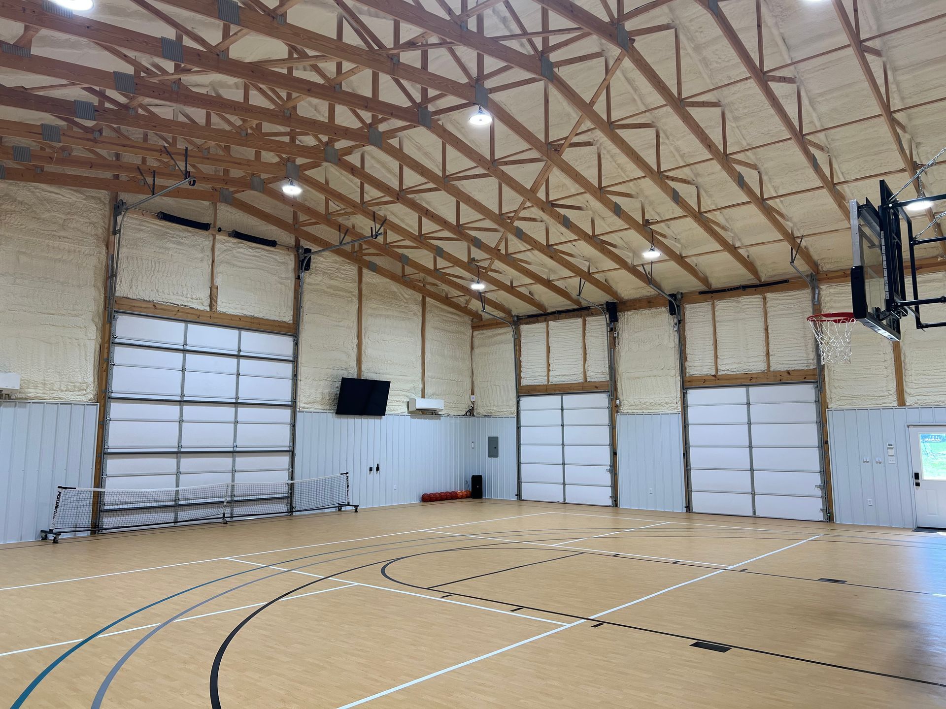 An indoor basketball court with a light-colored wood floor, light-colored walls, and a large wooden truss ceiling.