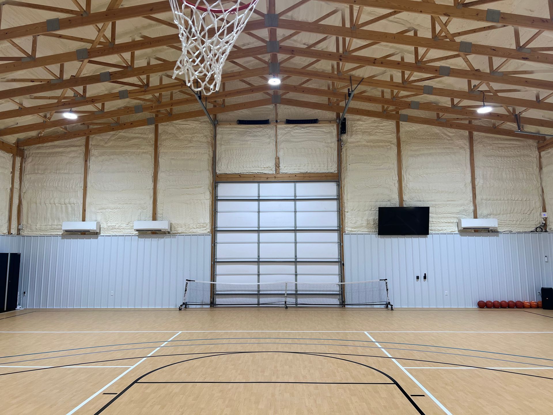 Indoor basketball court with light wood flooring, foam-insulated walls, a hoop, and a large central garage door.